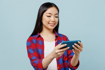 Young gambling woman of Asian ethnicity wear checkered shirt using play racing app on mobile cell phone hold gadget smartphone for pc video games isolated on plain pastel light blue color background