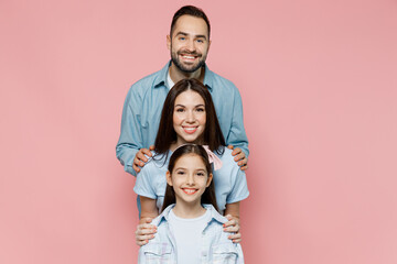 Young smiling cheerful happy parents mom dad with child kid daughter teen girl in blue clothes stand behind each other look camera isolated on plain pastel light pink background. Family day concept.