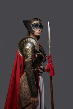 Studio shot of tattooed ancient amazon dressed in armor and red cloak holding spear.