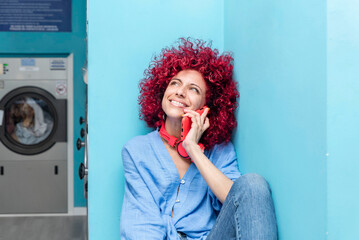 portrait of a smiling young latina woman with red afro hair sitting in a blue laundry room talking...