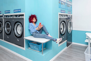 a bored young latin woman with a red afro hair sitting on a shelf in a blue laundry room eating a chupa chups listening to music and looking at her cell phone while waiting for the laundry to be done