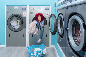 A smiling young latin woman with afro hair sitting inside a washing machine in a blue automatic laundry room