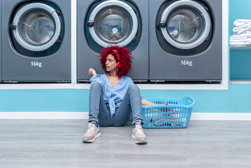 A smiling young Latina woman with red afro hair sitting in a blue automatic laundry room listening to music with red headphones while she waits for the laundry to be done.