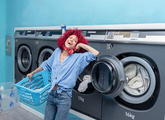 A laughing young latina woman with red afro hair listening to music while doing laundry in a blue automatic laundry room