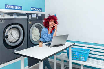 a smiling young latina woman with a red afro hair works with her laptop and talks on the phone in the blue laundry room, while waiting for the laundry to be done, her red headphones around her neck