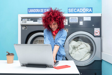 a smiling young latin woman with red afro hair works with her laptop in the blue laundry room while waiting for the laundry to be done, her red headphones around her neck