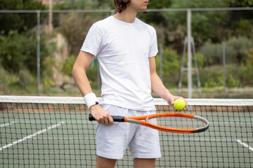 Close-up impersonal portrait of a tennis player holding the ball and a racket by the net on a green tennis court.