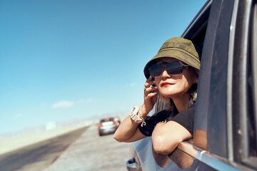 asian woman traveler with hat and sunglasses sticking head out of rear window of car looking at view