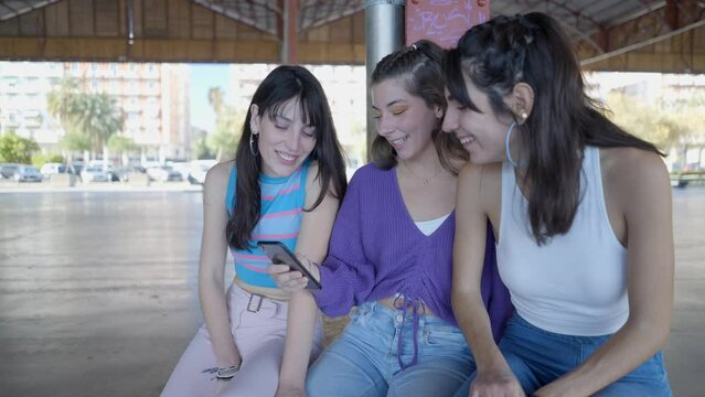 Three dark-haired teenage girls sit, look at phone and laugh outdoors