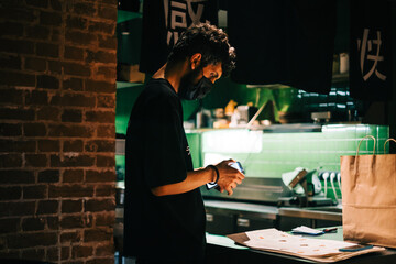 Young caucasian man asian bistro worker using mobile phone accepts online order for delivery or checks the order of guests in the hall.