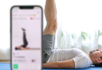 Home exercise using fitness app concept, young man works out, doing leg raises on blue yoga mat, following fitness app, white curtain in background covering houseplants, selective focus