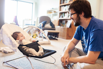 Father interacting with newborn baby in bouncer with messy house in background