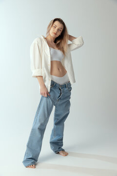 Full-length portrait of young slim girl wearing shirt, underwear and jeans isolated over grey studio background. Youth beauty