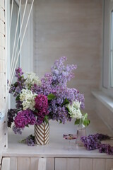 Fototapeta na wymiar A large lush bouquet of lilacs of different varieties in a vase on a bright loggia. Calm rest at home with a glass of soda.