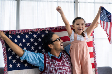 Naklejka premium Father and daughter raise and holding American flag and celebrate United State independence date inside of the living room. Shoe feeling of happiness and joyful. Family holiday event.