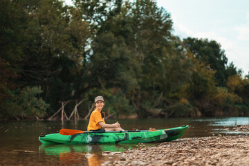 A young happy woman is sitting relaxed in a green kayak on the river bank. Copy space. The concept of kayaking and World Tourism Day