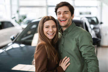 Portrait of happy young Caucasian couple posing and smiling at camera near new auto at dealership