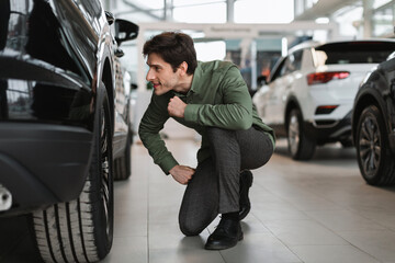 Positive young guy checking wheels of new car, buying modern vehicle at automobile dealership center
