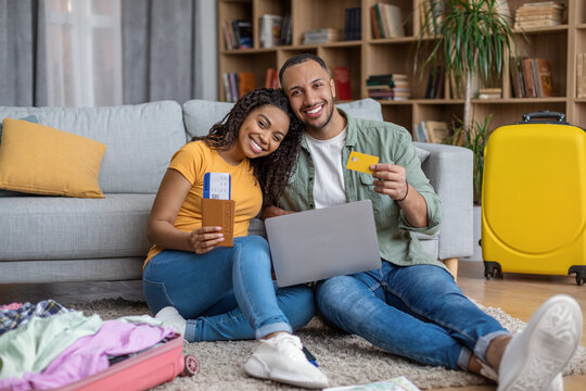 Happy african american spouses using laptop computer and buying travel tickets, sitting on floor with unpacked suitcases - Powered by Adobe
