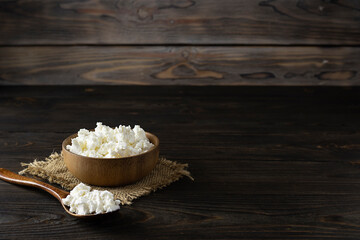 Fresh cottage cheese in a wooden bowl on a canvas napkin. Wooden spoon with cottage cheese on a dark table. Light brown wooden background, space for text