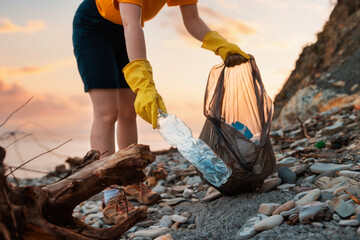 Ocean's pollutions. Close up of volunteer's hands in rubber gloves puts a dirty plastic bottle in bag. Sunset on the background. Bottom view. The concept of ecology disaster