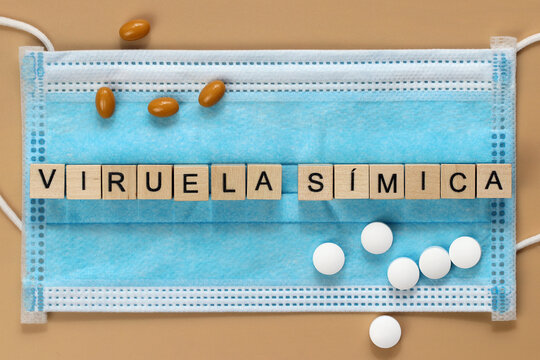 Viruela s&iacute;mica is the name of the monkeypox virus in Spanish. The word is laid out with wooden cubes on a surgical face mask. There are various pills lying around.