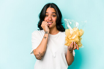 Young hispanic woman holding a bag of chips isolated on blue background biting fingernails, nervous and very anxious.