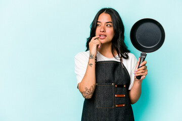 Young hispanic cooker woman holding frying pan isolated on blue background relaxed thinking about...