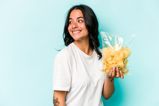 Young hispanic woman holding a bag of chips isolated on blue background looks aside smiling, cheerful and pleasant.