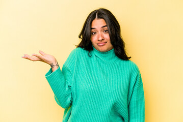 Young hispanic woman isolated on yellow background doubting and shrugging shoulders in questioning gesture.