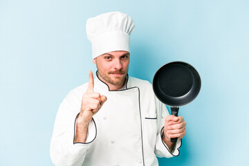 Young caucasian chef man holding flying pan isolated on blue background having an idea, inspiration concept.