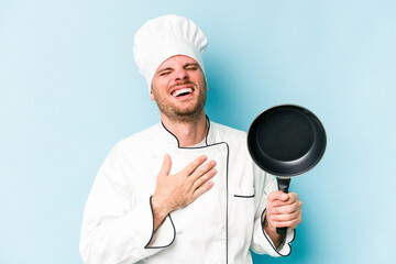 Young caucasian chef man holding flying pan isolated on blue background laughs out loudly keeping hand on chest.