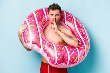 Young caucasian man holding an inflatable donut isolated on blue background keeping a secret or...