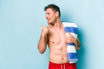 Young caucasian man holding beach towel isolated on blue background points with thumb finger away,...