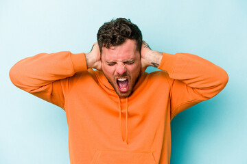 Young caucasian man isolated on blue background covering ears with hands trying not to hear too loud sound.