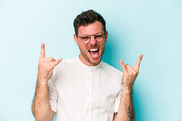 Young caucasian man isolated on blue background showing a horns gesture as a revolution concept.