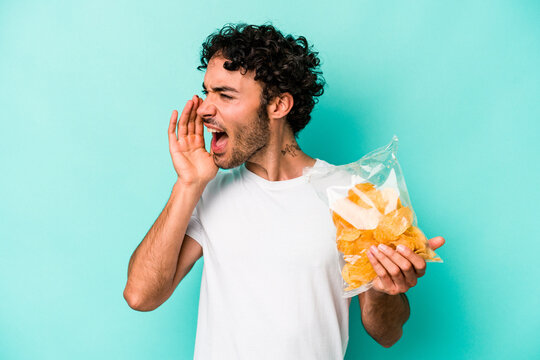 Young Caucasian Man Holding A Bag Of Chips Isolated On Blue Background Shouting And Holding Palm Near Opened Mouth.