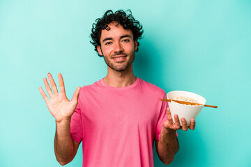 Young caucasian man eating noodles isolated on blue background smiling cheerful showing number five...