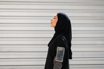 Tired young arab woman in hijab with phone on shoulder resting after exercise on gray wall background, profile