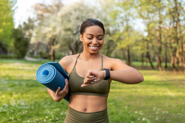 Happy black woman looking at smartwatch, tracking fitness activity, holding sports mat, ready for training outdoors