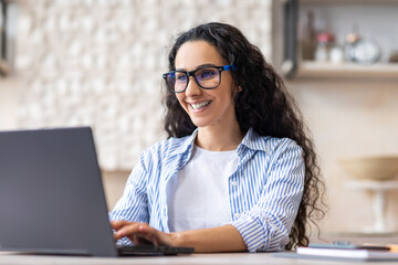 Positive latin female freelancer in eyeglasses using laptop at home, sitting at table in kitchen, typing on keyboard