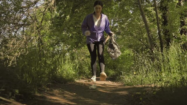 A young woman runs through the forest and collects garbage in the forest. Low angle view. Follow shot. The concept of plogging and volunteering.