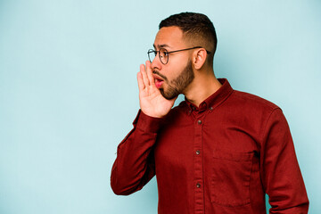 Young hispanic man isolated on blue background is saying a secret hot braking news and looking aside