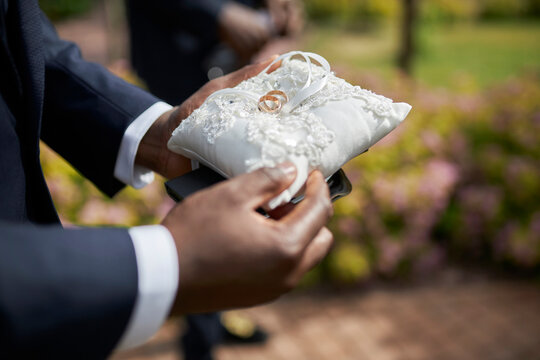 An African American Groom's Friend Holds A White Pillow With Wedding Rings In His Hands. Close-up Of African American Man's Hands With White Pillow With Wedding Rings