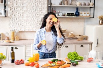 Happy young latin lady having fun and covering eyes with lemon halves, fooling around in kitchen while cooking dinner