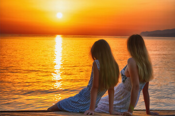 Mom and daughter sitting on a bridge by the sea and meeting morning sunrise. Summer holiday, rest,...