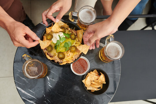 Hands of people drinking beers with eating bowl of nacho chips with rice, jalapeno slices, guacamole and sour cream, view from above
