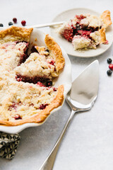 A berry pie with two slices taken out. One slice is still in the pie pan and the other is on a plate to the side. There is a pie serving utensil and berries all around. A delicious treat.