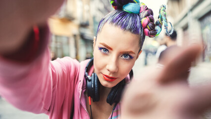 Playful cool funky hipster young girl with headphones and crazy hair taking selfie on street