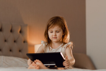 Cute toddler little girl wearing casual dress sitting lying in bedroom with modern interior in beige colors using laptop tablet pad.